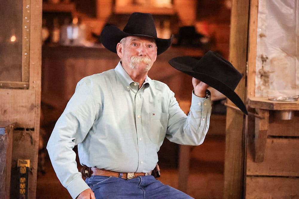 Un vaquero con bigote gris, que lleva un sombrero de vaquero negro y sostiene un sombrero de vaquero negro.